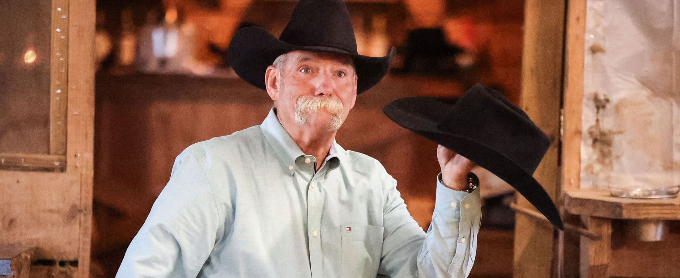 Un vaquero con bigote gris, que lleva un sombrero de vaquero negro y sostiene un sombrero de vaquero negro.